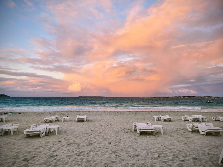 Beach with white chairs facing the ocean. The sky is pink from the sunset.